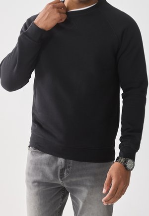 Black sweatshirt in soft fabric with a round neckline and ribbed cuffs, paired with grey jeans and a silver watch.
