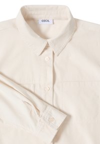 Camicia beige con colletto realizzata in tessuto testurizzato, dotata di due tasche anteriori, chiusura con bottoni e un'etichetta con "CECIL" sul colletto.