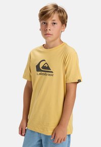 Žluté bavlněné tričko s krátkým rukávem, na přední straně s černým logem Quiksilver a grafikou hory, hladká textura.