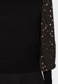 Maglione nero con corpo aderente e maniche larghe a fantasia. Le maniche presentano pois dorati su un tessuto scuro, che aggiungono texture e dettaglio.