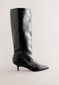 Botte en cuir noire mi-mollet avec un bout pointu, un petit talon kitten et un motif texturé subtil sur un fond neutre.