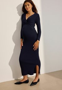 Marineblaues, geripptes Schwangerschafts Kleid mit gedrehtem Vorderteil, langen Ärmeln, seitlichem Schlitz und taillierter Silhouette, kombiniert mit schwarzen spitzen Ballerinas.