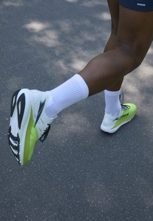 White and neon green running shoe with a black sole, featuring a mesh upper and laces, worn with white athletic socks on a pavement background.