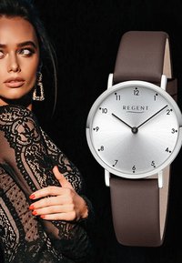 Montre ronde argentée avec un cadran brossé, index horaires noirs et un bracelet en cuir brun. Présente un design minimaliste avec une bonne visibilité.