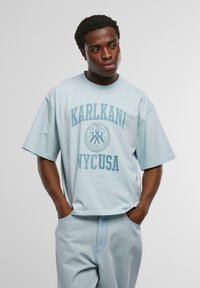 Tricou oversize de culoare albastru deschis, cu mâneci scurte, având un grafic central care spune "KARLKANI NYCUSA" în nuanță de albastru mai închis. Design simplu, cu o croială relaxată.