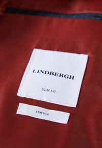 Vita tygetiketter med svart text som säger "LINDENBERGH" och "SLIM FIT" på en röd satängbakgrund med en mindre etikett under som säger "STRETCH".