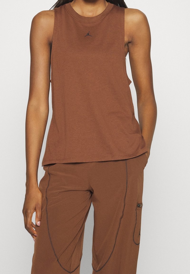 Femme portant un haut marron sans manches et un pantalon marron assorti avec des détails de couture noire et un petit logo noir sur le devant du haut.