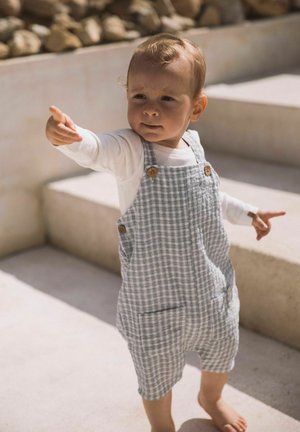 Vauva, jolla on harmaat ruudulliset haalari ja valkoinen pitkähiha-paita, ojentaa kättään seisoessaan ulkoportailla auringonvalossa.