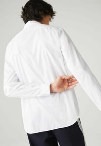 Witte lange mouwen shirt met knoopmanchetten en een gebogen zoom, gemaakt van een gladde stof. De hand van het onderwerp reikt naar achteren en verbergt gedeeltelijk de details van het shirt.
