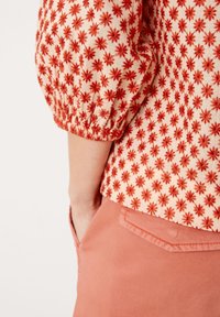 Rød bluse med blomsterbroderi og pufærmer, der har en cremefarvet baggrund. Ved siden af let koralfarvede bukser med detalje af en baglomme.