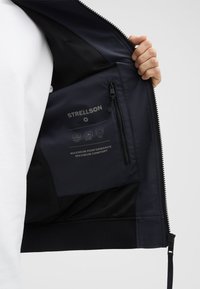 Navy Zip-Jacke mit Mesh-Futter, ausgestattet mit einer Innentasche mit STRELLSON-Branding und Pflegeanweisungen. Der Text hebt Komfort und Leistung hervor.