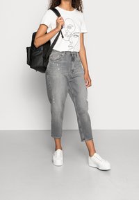 Biała koszulka z rysunkami twarzy, szare zniszczone jeansy typu mom z skróconym dołem oraz białe trampki na platformie; czarna plecak na ramieniu.