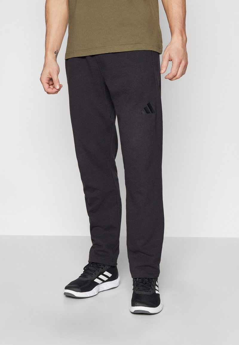 Juodi medvilniniai džinsai turi tekstūruotą apdailą, elastingą juosmenį ir mažą juodą Adidas logotipą ant kairiojo šlaunies. Su jais derinami juodi sportiniai batai.