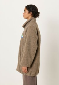 Sani ruskea fleece-takki, jossa on korkea kaulus, ylisuurta muotoilua ja rintapielessä kuvakkeita ja tekstiä sisältävä kangasmerkki. Pehmeä tekstuuri ja rento muotoilu.