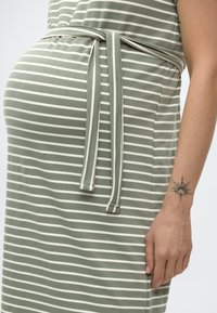 Έγκυος γυναίκα που φορά ένα πράσινο και άσπρο ριγέ φόρεμα με δεμένη ζώνη και μαύρο τατουάζ ήλιου στον αριστερό καρπό της.