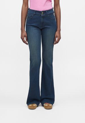 Femme portant un jean évasé bleu foncé, un haut rose rentré et des baskets beiges, debout, les bras détendus le long du corps.