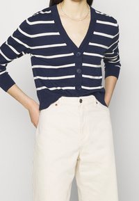 Granatowy i biały sweter w paski z dekoltem w kształcie V, z pięcioma guzikami, długimi rękawami i dopasowanym fasonem, zestawiony z kremowymi spodniami.