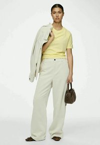 Woman wearing yellow t-shirt and white wide-leg pants, holding matching white jacket over shoulder and brown handbag, leopard print shoes.