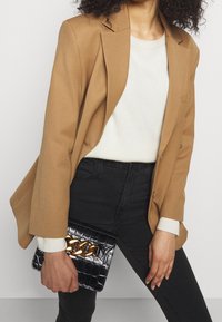 Blazer beige, suéter blanco, jeans negros; sosteniendo un bolso de charol negro con un detalle de cadena dorada. Tela suave con forma estructurada.