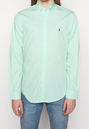 Homme portant une chemise à rayures verticales vert clair et blanc avec un logo brodé violet sur la poitrine, associée à un jean bleu.