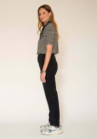 Chemise cropped à rayures noires et blanches, pantalon en velours côtelé noir et baskets argentées. Cheveux longs et ondulés, portant des bijoux. Fond uni.