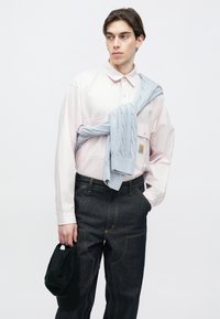 Hele roosa nööpidega särk, tumedad teksapüksid kollase õmblusega ja hele sinine soonikkoes kampsun õlgadel seotud. Must nokamüts käes.