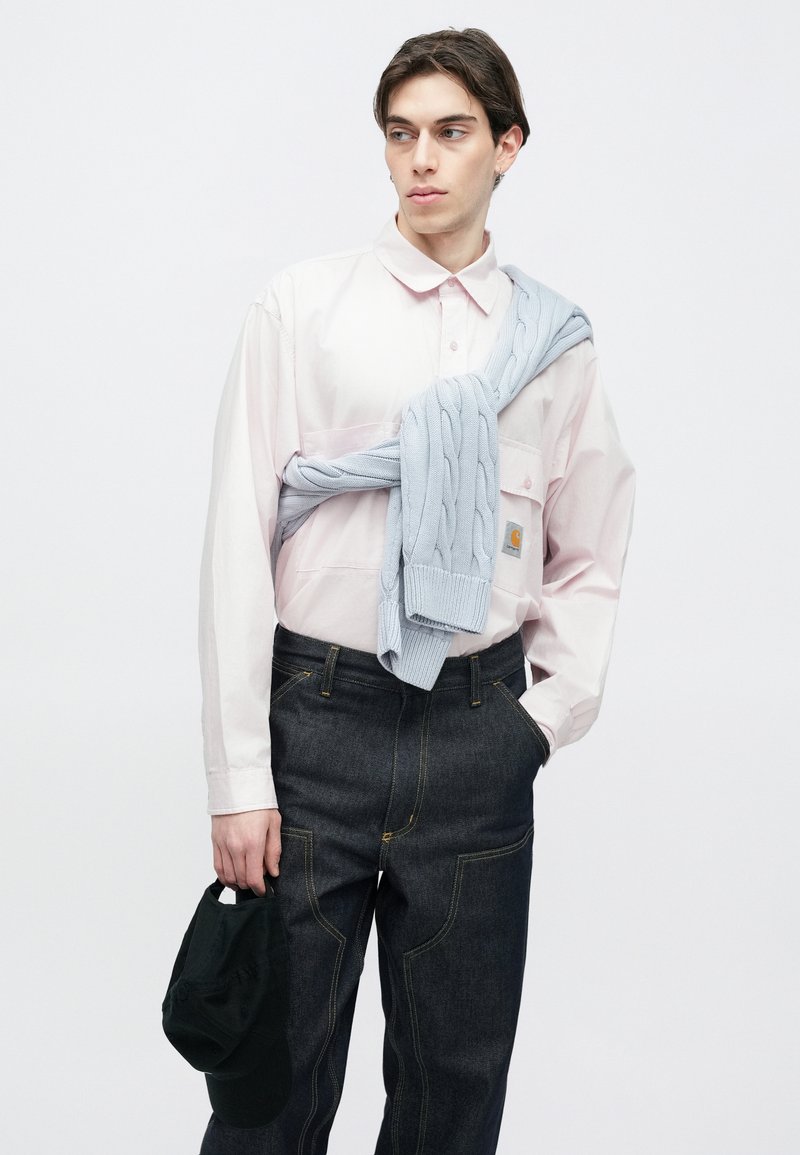 Ljusrosa skjorta med knappar, mörka jeans med gula sömmar och en ljusblå kabelstickad tröja knuten runt axlarna. Svart keps i handen.