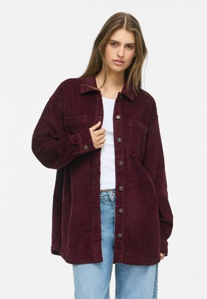 Jovem com cabelo comprido a usar uma blusa de veludo cotelê larga de cor vermelho escuro com botões, por cima de um top branco e jeans azuis, contra um fundo simples.