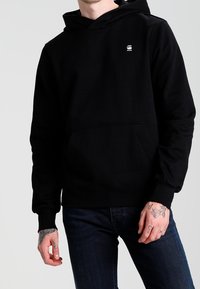Sudadera negra con bolsillo frontal y un pequeño logo en el pecho. Fabricada con tejido suave, cuenta con capucha ajustable y puños acanalados.