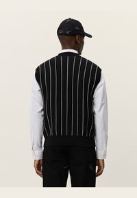 Svart strikket vest med hvite vertikale striper, med ribbestrikket mønster i kanten og rund hals, båret over en hvit langermet skjorte.