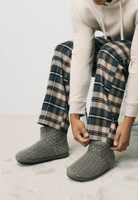 Grå teksturerede tøfler med et zigzag-mønster, i midt-læghøjde, gummisåler, båret med ternede hjemmetøfler i blå og beige nuancer.