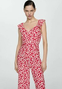 Žena v květinovém overal s bezrukávovými červenými a bílými motivy a volánkovými detaily na ramenou, stojící na jednobarevném světlém pozadí.