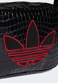 Črna torbica z reliefnim vzorcem krokodila in rdečim izvezenim logotipom s tremi listi ter vodoravnimi črtami. Gladek teksturni videz in zadrga za zapiranje.