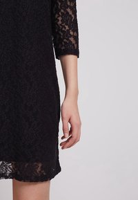 Svart spetsklänning med trekvartsärmar, med blommönster och en fåll med intrikata vågkanter, delvis synlig.