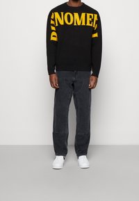 Pull en maille noire avec grand texte jaune "DIE NOMMEE". Associé à un pantalon gris foncé et des baskets blanches. Tenue simple et décontractée.