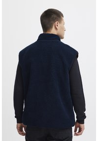 Tamno plavi polu-dukserica bez rukava s visokim ovratnikom, koja ima teksturiranu površinu i ležeran kroj, nosi se preko teksturirane crne majice s dugim rukavima.