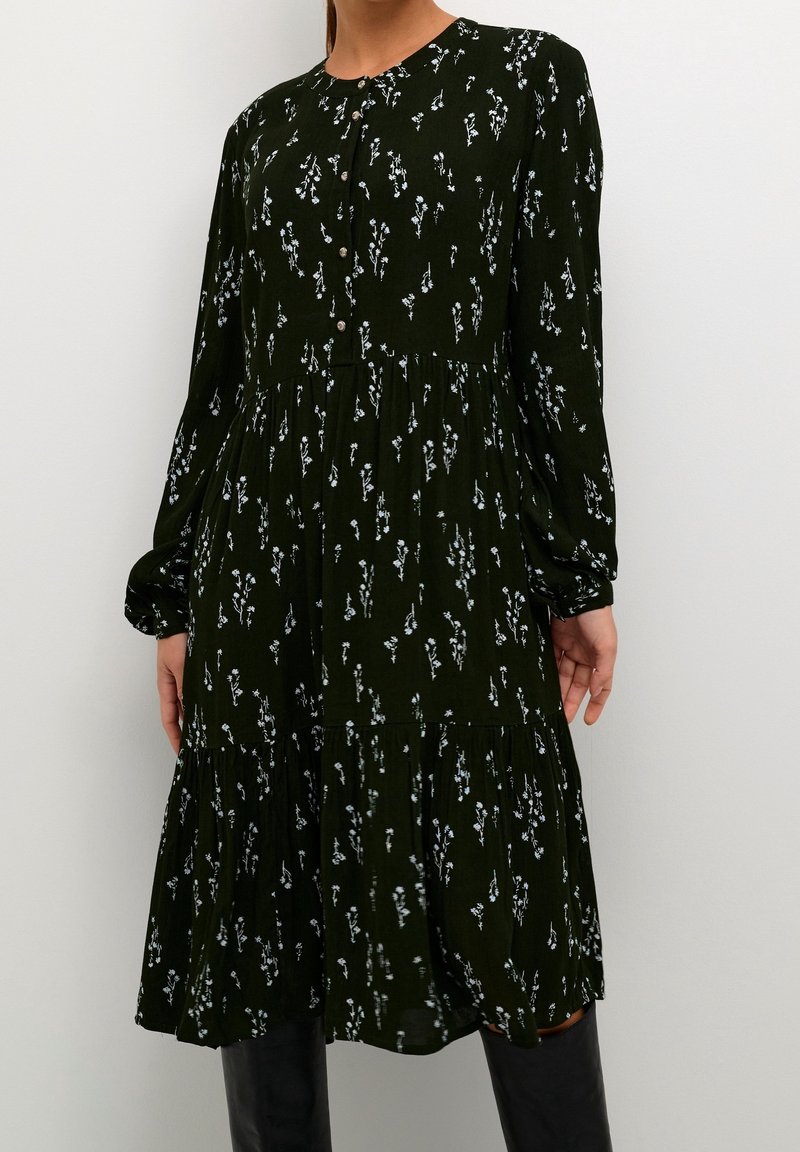 Robe midi noire à manches longues avec petit imprimé floral blanc, boutonnée devant, taille froncée et jupe légèrement évasée portée avec des bottes noires.