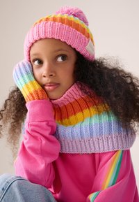 Cappello lavorato a maglia con strisce multicolori e pon pon; scaldacollo a righe abbinato; maglione rosa; capelli ricci; posa rilassata.