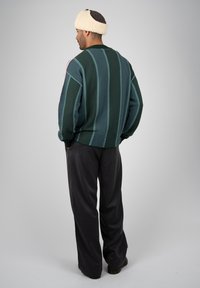 Homme portant un pull rayé vert et bleu, un pantalon foncé et un chapeau en laine crème, debout, les mains dans les poches, tourné de dos.