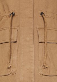 Tan jakke med snorelukning, to forlommer, tekstureret stof og synlige syninger. Funktionelt design med holdbare egenskaber.