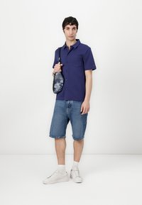 Koszula z krótkim rękawem w granatowym kolorze, z guzikami i kieszonką, połączona z dżinsowymi szortami. Model trzyma czarną torbę w wzór tie-dye i ma na sobie białe trampki.