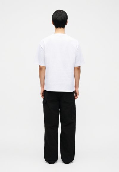 Άτομο που φαίνεται από πίσω, φοράει απλό λευκό μπλουζάκι και φαρδιά μαύρη παντελόνα cargo, με φωτεινό και απλό φόντο.