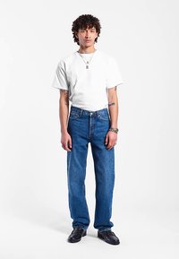 Bílý tričko s krátkým rukávem v kombinaci s modrými džínami s rovnými nohavicemi. Model nosí černé boty a doplňky zahrnují náhrdelníky a stříbrný náramek.