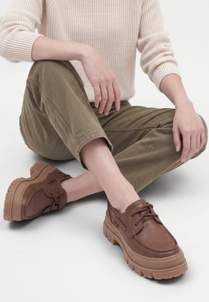 Person, der sidder med benene over kors iført en cremefarvet ribstrikket sweater, olivengrønne bukser og brune lædersko med tyk sål på en hvid baggrund.