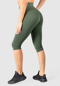 Leggings a vita alta di colore verde oliva realizzati in tessuto elasticizzato, con lunghezza cropped e superficie liscia, abbinati a scarpe da ginnastica nere.