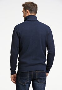 Marinblå, stickad tröja med hög krage, strukturerat mönster och ribbade ärmslut. Kombinerad med mörka jeans, som visar bakvyn.