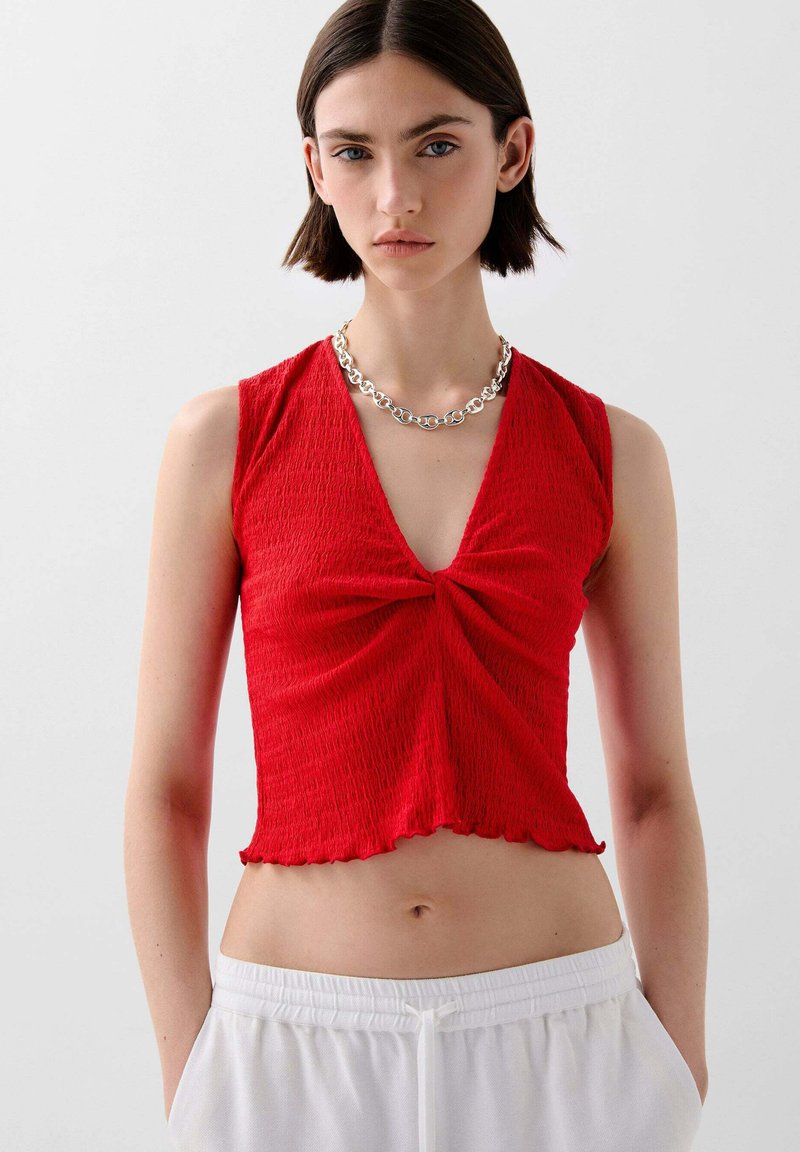 Rotes ärmelloses Top mit tiefem V-Ausschnitt und gedrehtem Frontdetail. Strukturstoff mit gewelltem Saum. Kombiniert mit weißen Jogginghosen mit Kordelzug.