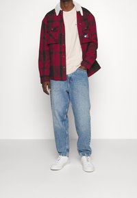 Röd och svart rutigt flanellskjorta med en vit sherpa-krage, lager på en beige t-shirt, tillsammans med ljusblå lösa jeans och vita sneakers.