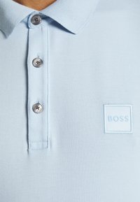 La polo di colore blu chiaro presenta un tessuto testurizzato, una chiusura a tre bottoni e un logo quadrato ricamato con la scritta "BOSS" sul petto.