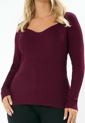Femme portant un pull ajusté en côtes à manches longues de couleur violet foncé avec un col arrondi, associé à un pantalon noir.